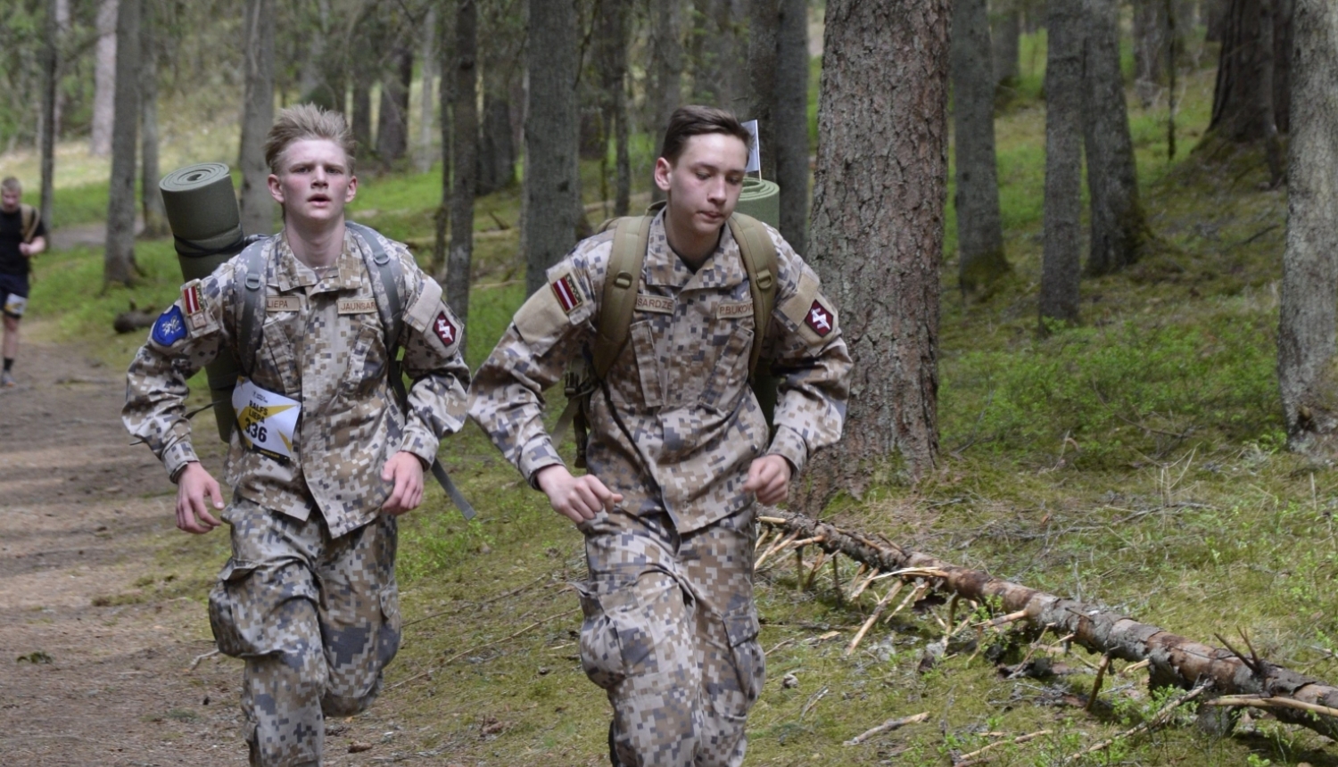 Rucking seriāla posms - jaunieši, tērpti militārajā apģērbā, skrien ar mugursomām
