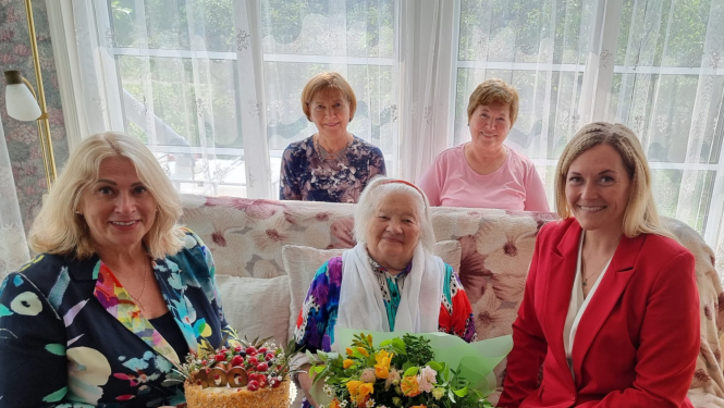 Centrā 100 gadu jubilāre ar ziediem un torti, apkārt 4 sievietes