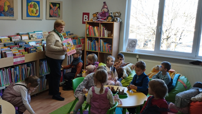 bērni bibliotēkā skatās grāmatas un klausās bibliotekāres stāstījumā