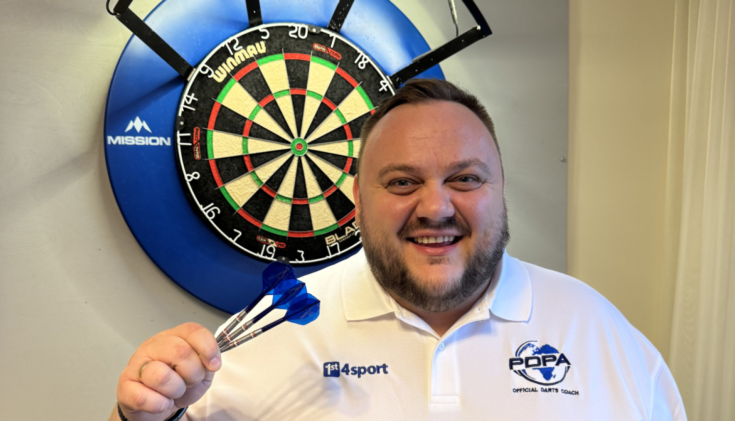 Jānis Dāmis tur rokā šautriņas, fonā šautriņu mešanas aplis