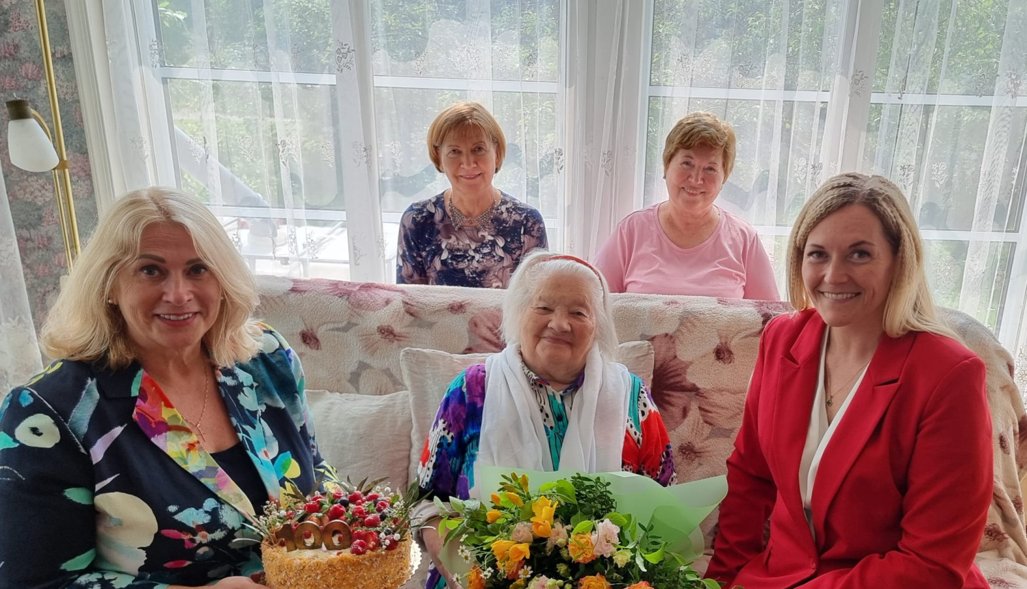 Centrā 100 gadu jubilāre ar ziediem un torti, apkārt 4 sievietes
