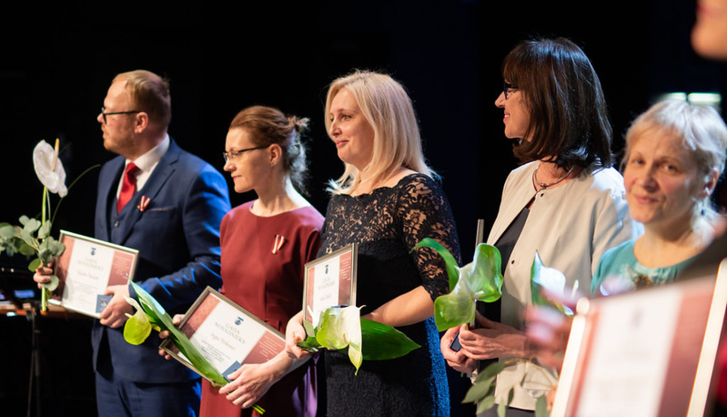 cilvēki ar ziediem un pateicības rakstiem uz skatuves