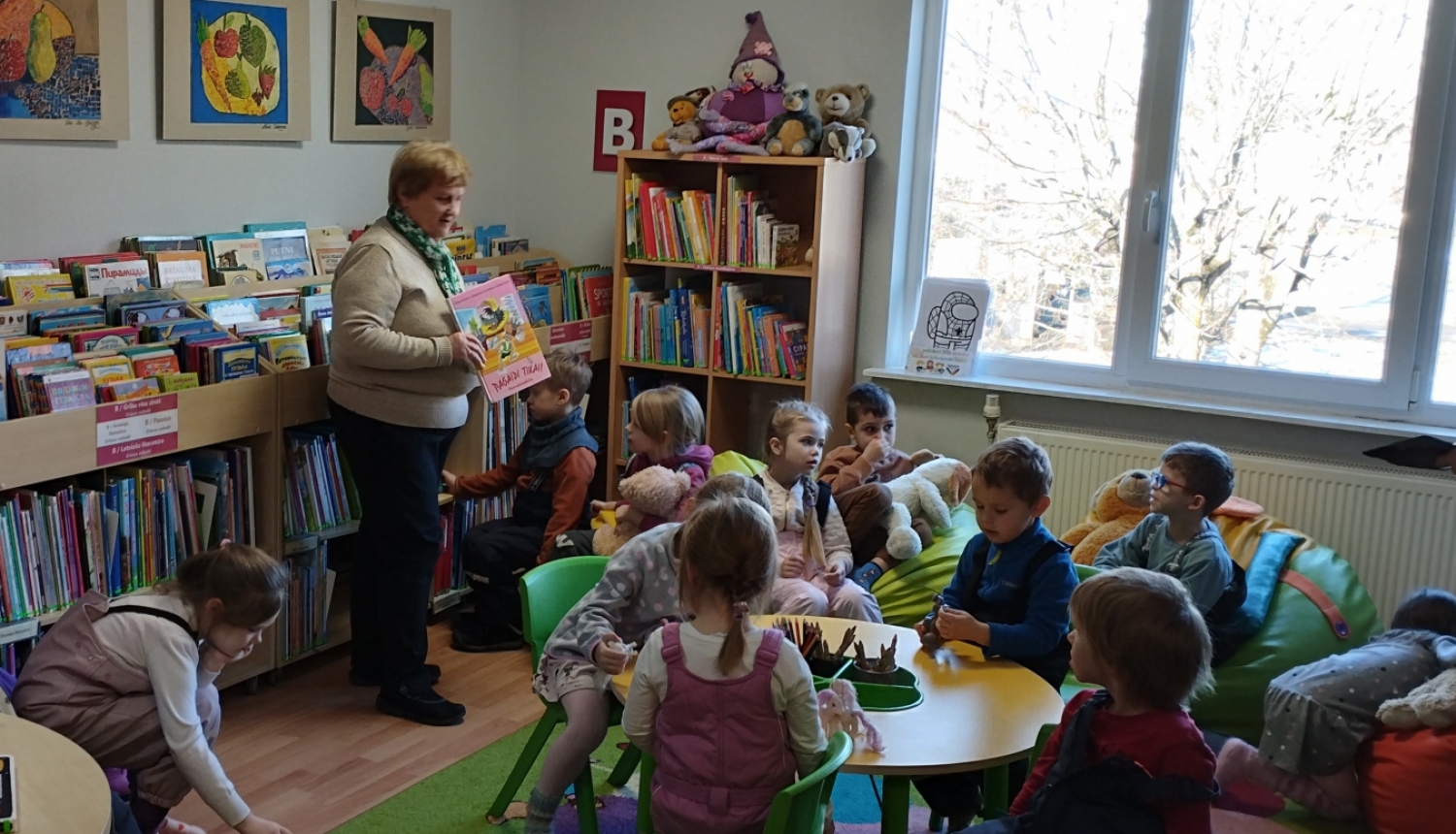 bērni bibliotēkā skatās grāmatas un klausās bibliotekāres stāstījumā